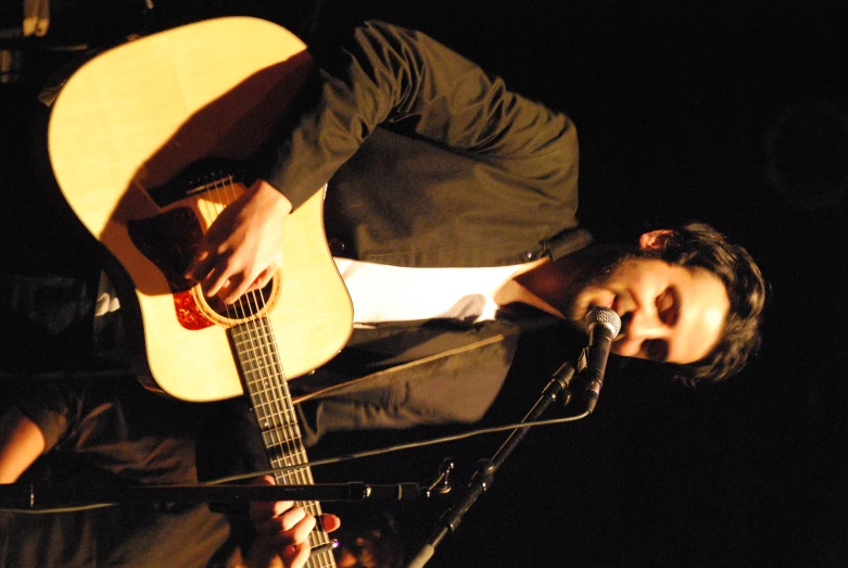man with long curly hair in a suit, playing an acoustic guitar