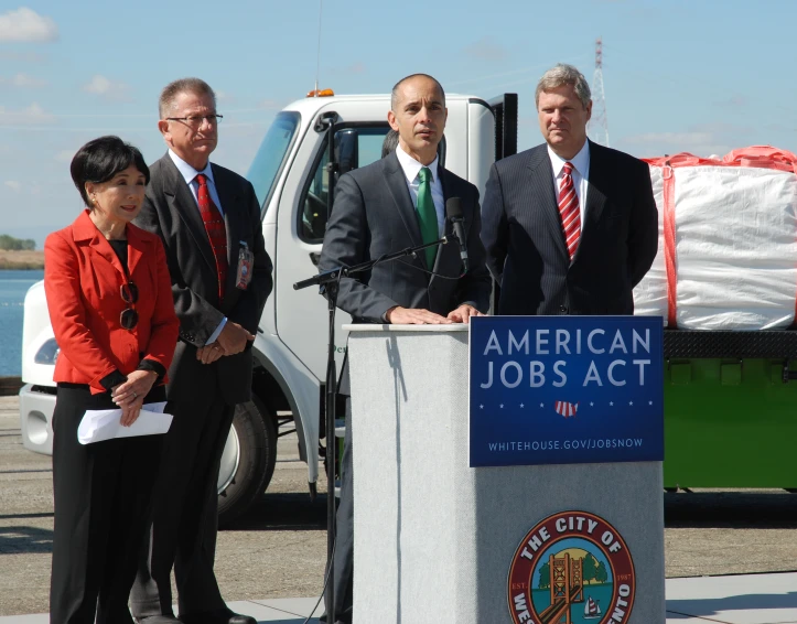 four people standing at a podium in front of an american job act truck