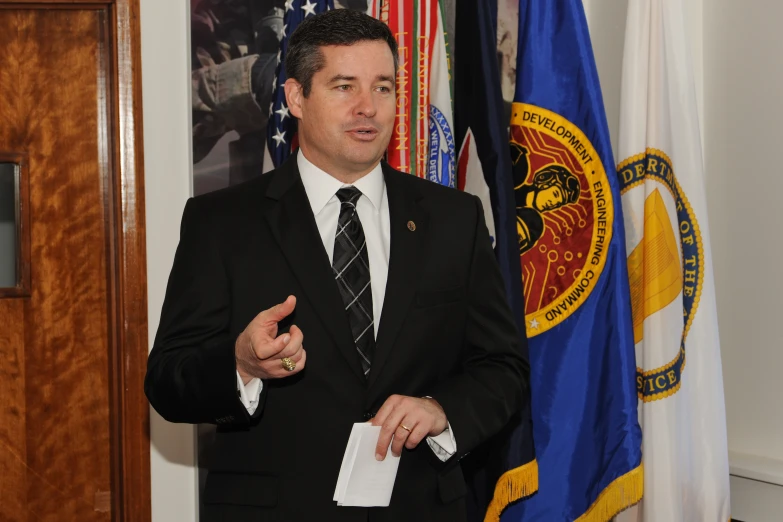 a man in a suit speaks with two flags behind him