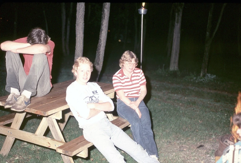 two young men sitting on picnic tables next to firewood