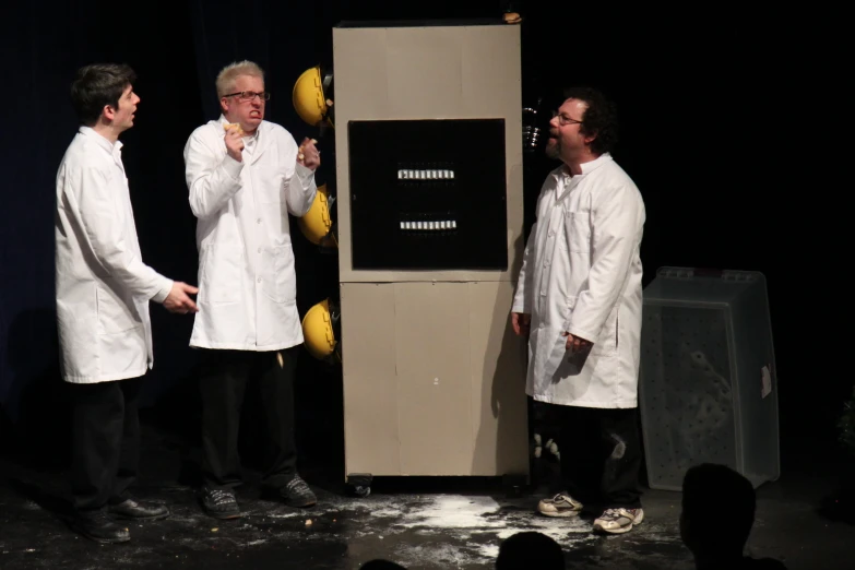 three men in white lab coats stand around a podium