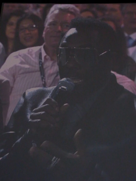 a man in a suit and sunglasses sits in front of a crowd