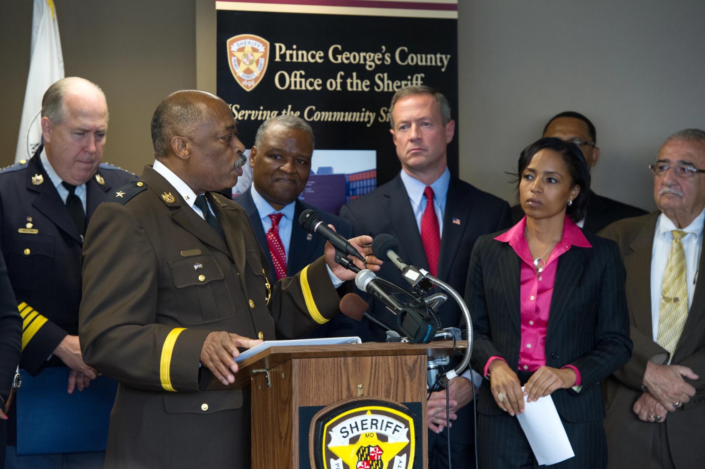 a man in uniform talking at a podium with officials standing behind him