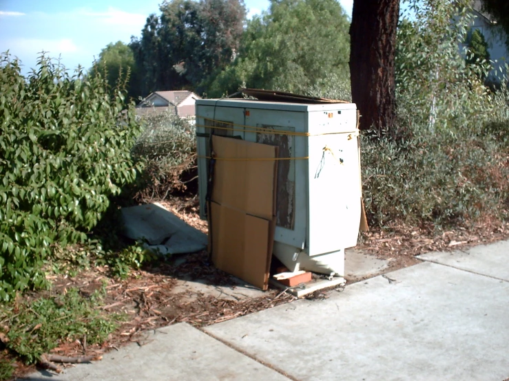 a broken refrigerator next to trees in a suburban area