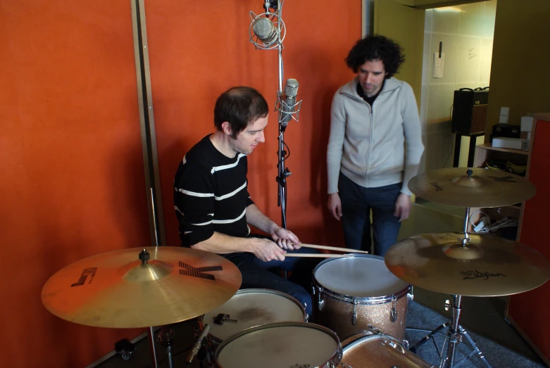 two men in an orange room, one is playing drums while the other is behind them