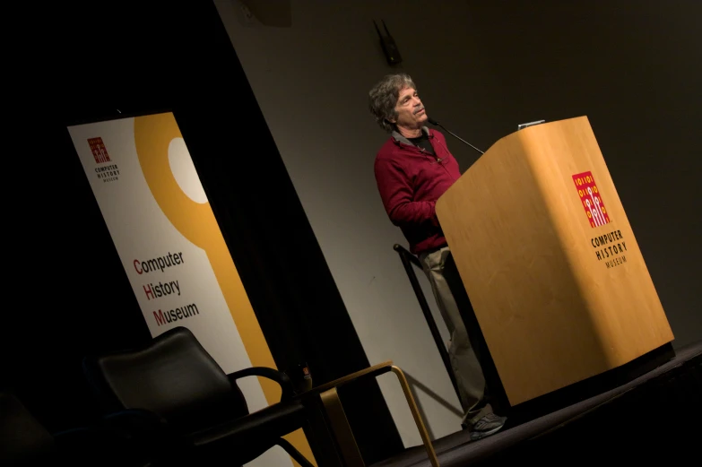 an old woman wearing a red jacket standing at a podium with a laptop