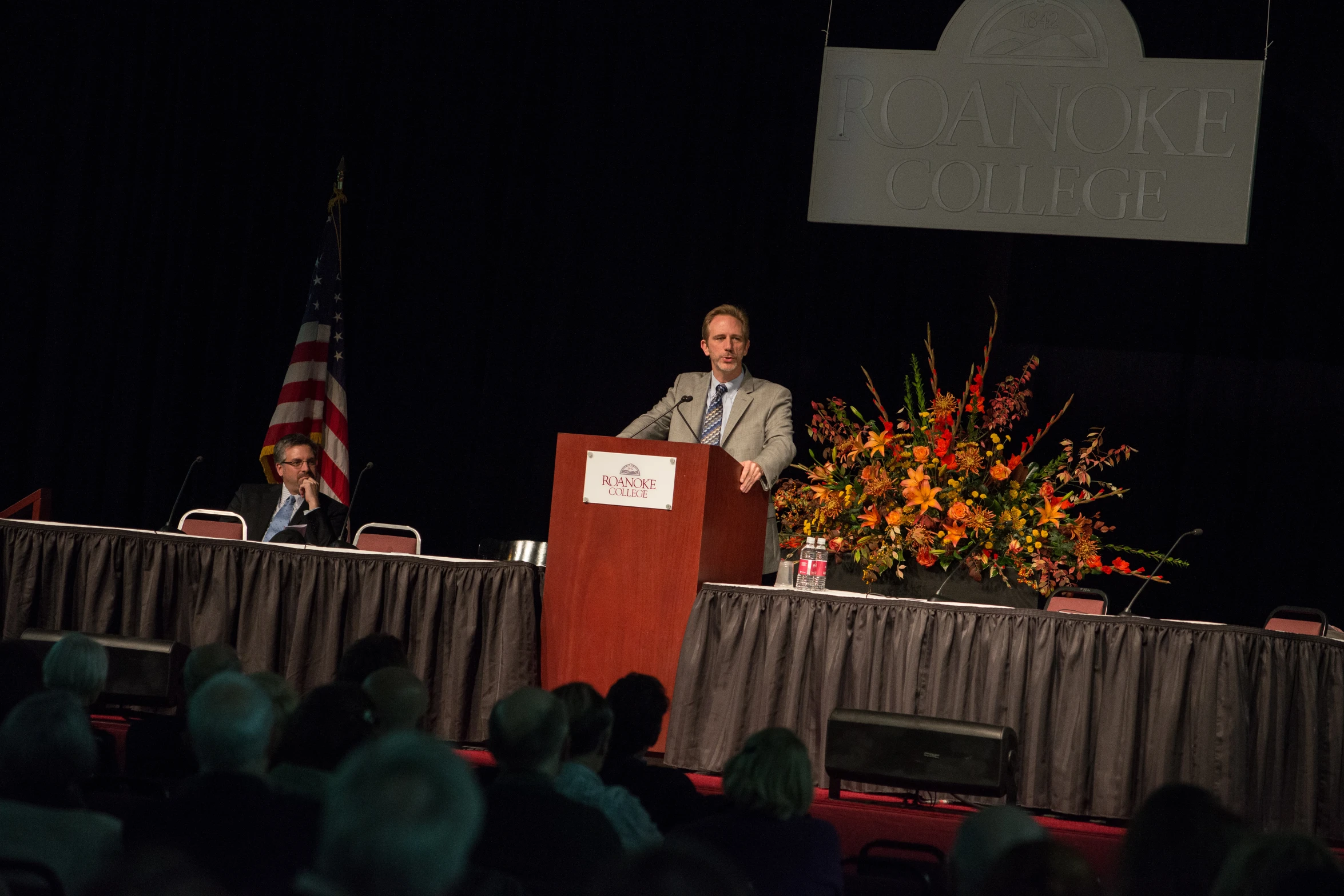 man in business attire standing at podium talking on a stage