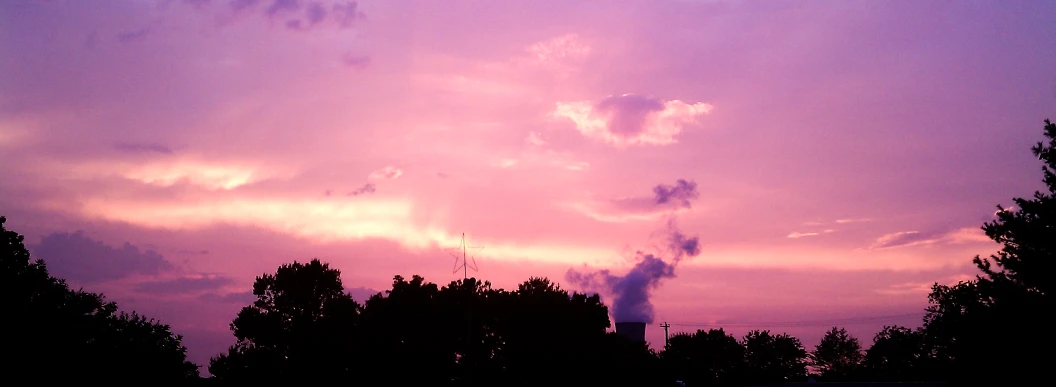 a purple sunset over some trees, with a plane in the sky