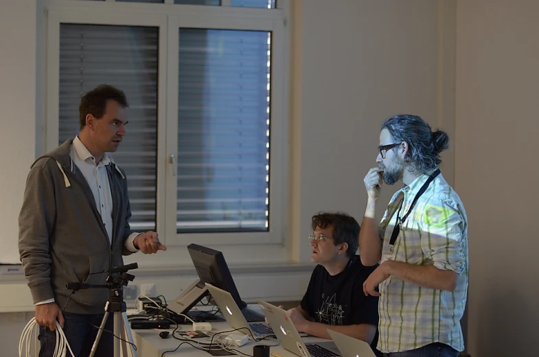 two men standing next to each other at a desk with three computers