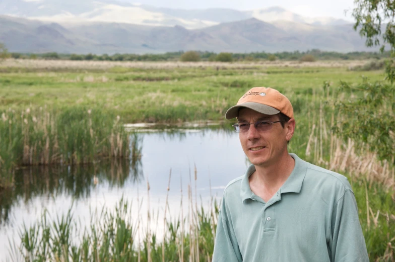 a man wearing a brown hat standing in a marsh with mountains in the background