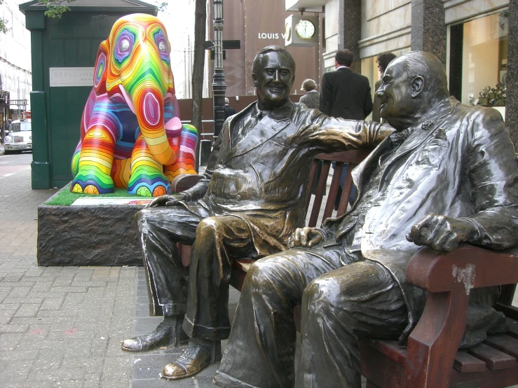 two statues of men sitting on benches near a rainbow elephant sculpture