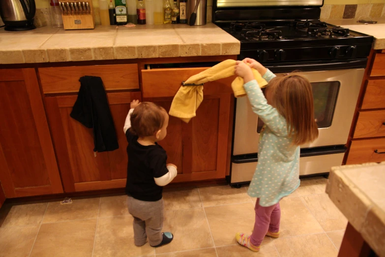 two children standing in front of a kitchen counter, putting cloth over the oven and holding on to a towel