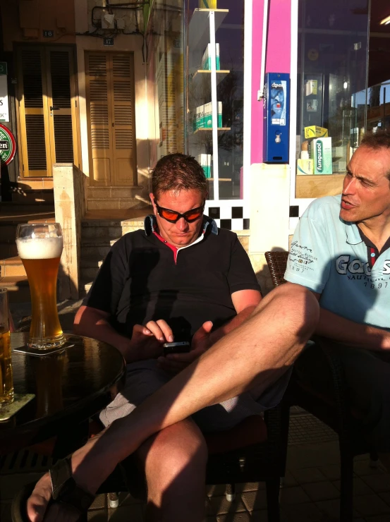 two men sit at a table outside with beer