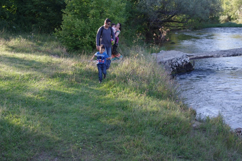 a couple of people walk down a grass covered path by a river