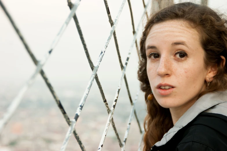 a girl with brown hair and wearing a gray shirt leans against a fence