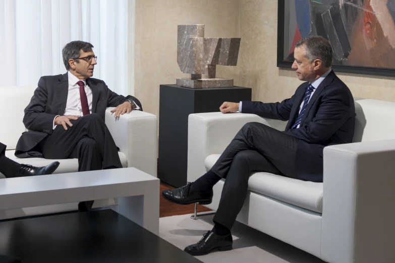 two men talking while sitting in chairs in the living room