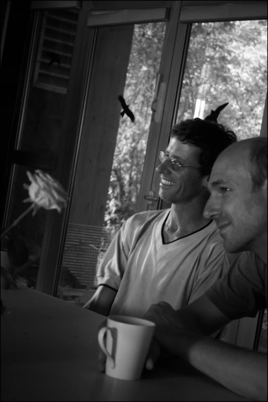 two men are sitting near each other on a table in front of windows