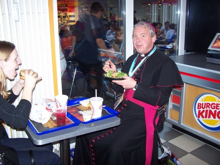 a man in priest clothing sitting at a diner with his drink and a woman standing near by
