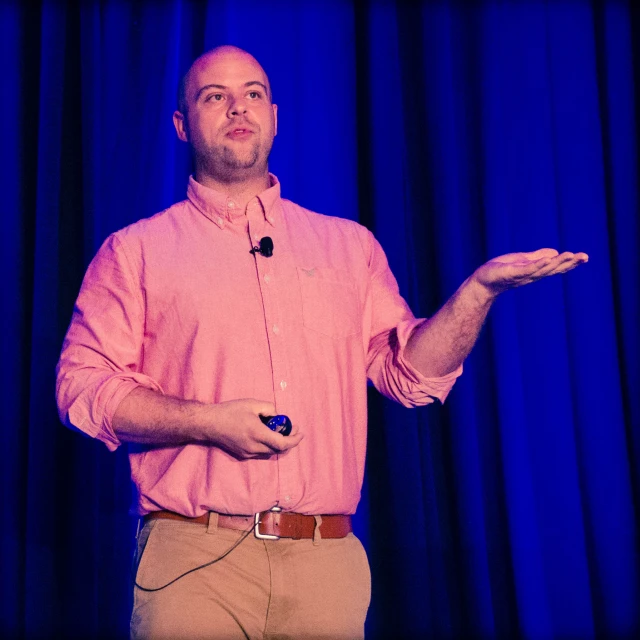 a man stands in front of blue curtain, holding a remote and gesturing at soing