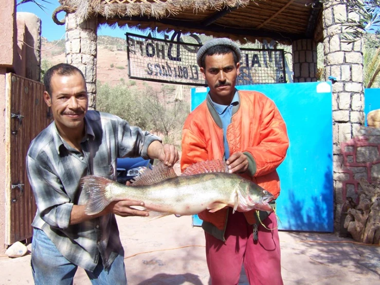 two men holding a fish standing outside of a building