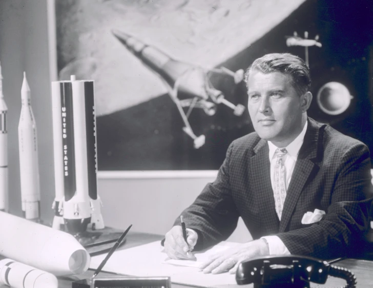 black and white po of man in suit sitting at desk with model airplane