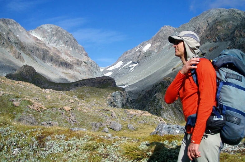 a man in an orange shirt and backpack walks along a field with snow covered mountains