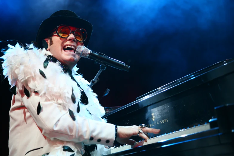 a man with glasses and a feather jacket playing a piano