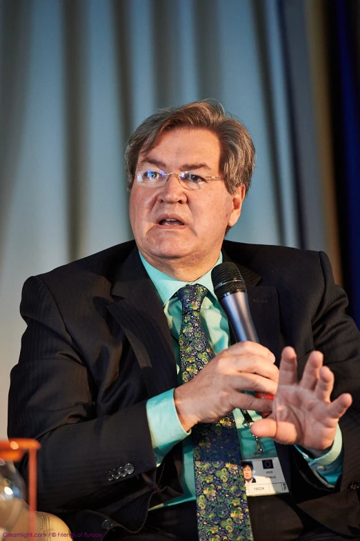 a man sitting and gesturing while talking into a microphone