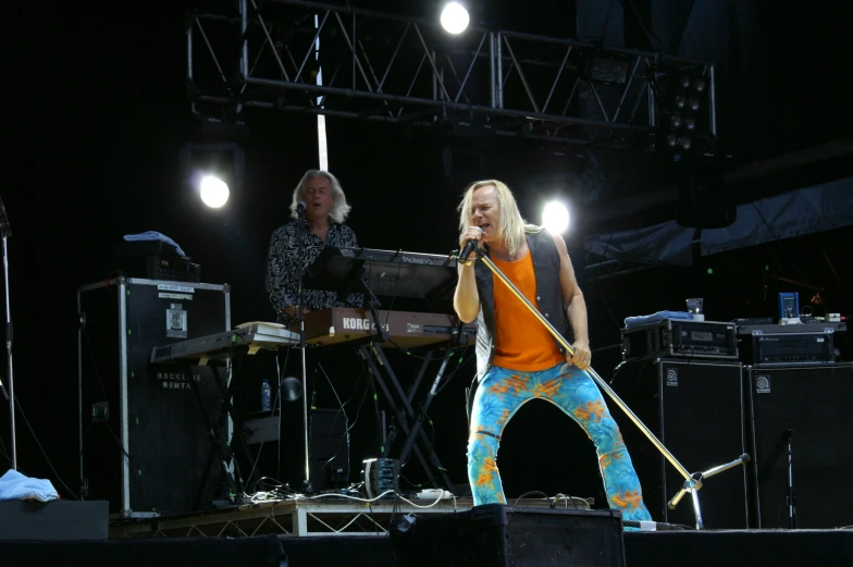 a female with long blonde hair and pants on singing on stage with two band members