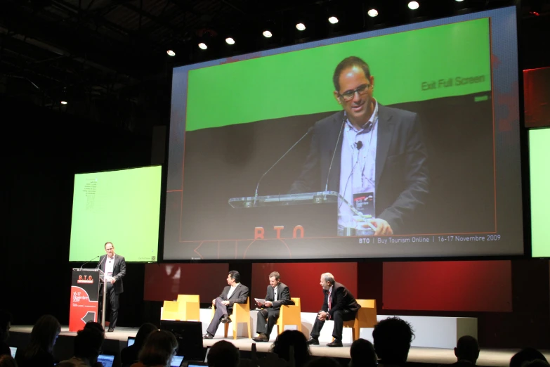 three men are on stage with video on the screen