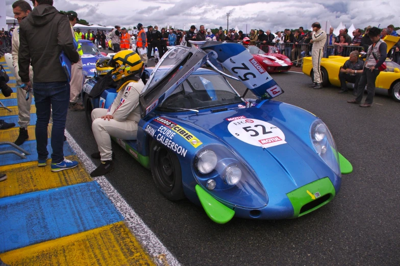 several people standing around a small racing car in a parking lot