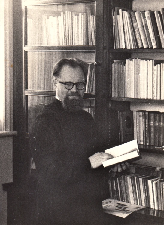 a man with beard holding an open book in front of a bookshelf