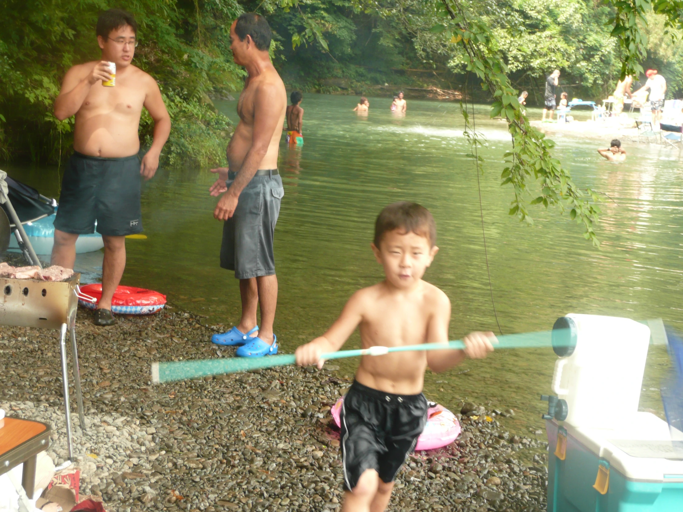 a boy holding a plastic object on the shore of a river while another  watches