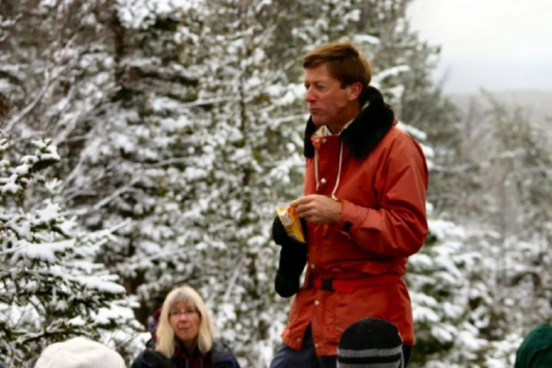 a man in a red jacket standing in front of snow covered trees and with a cigarette in his mouth