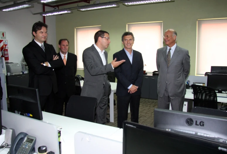 four men are standing in an office with computer monitors and monitors