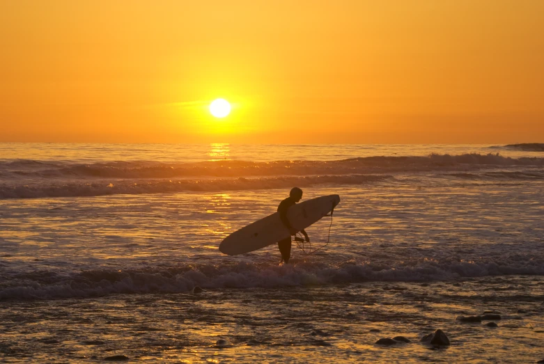 a man carrying a surfboard standing in the ocean at sunset