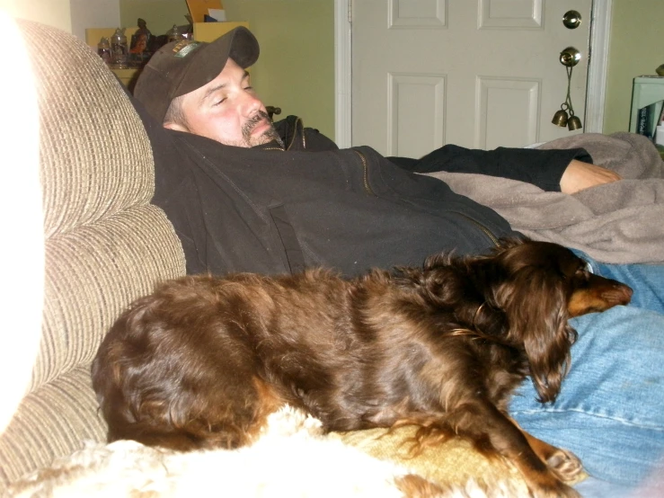 a man in black jacket and cap laying on a couch with brown dog