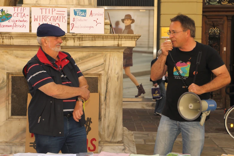 two men are standing by a stone fireplace with papers, and one holds a megaphone