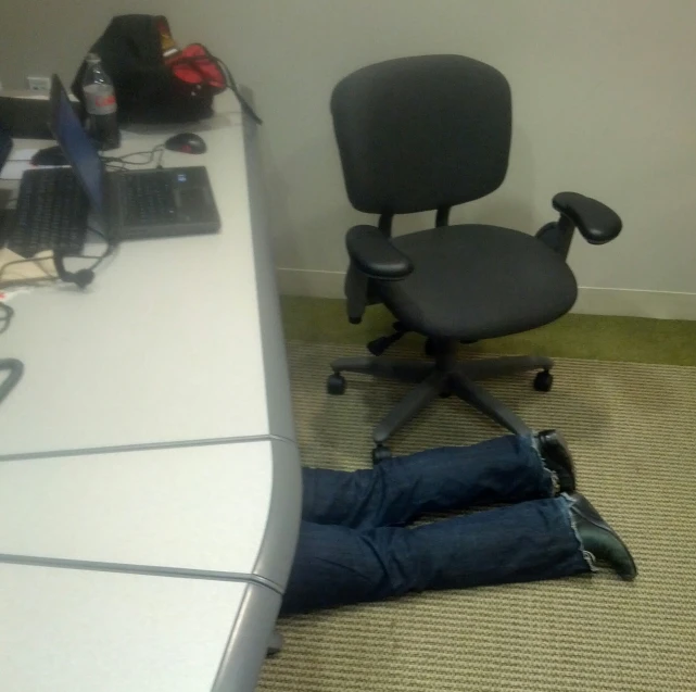 a man with blue jeans laying on the floor under a computer desk