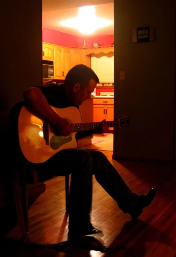 man sitting on chair holding guitar with bright sunlight streaming through room