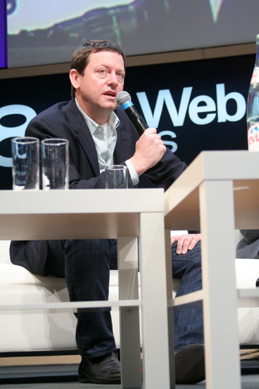 man in black blazer sitting on a white couch while talking into microphone