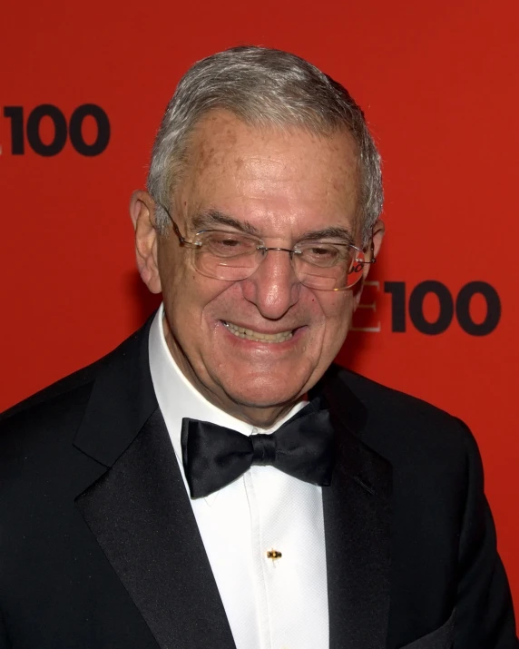 an older man in glasses and a suit stands smiling
