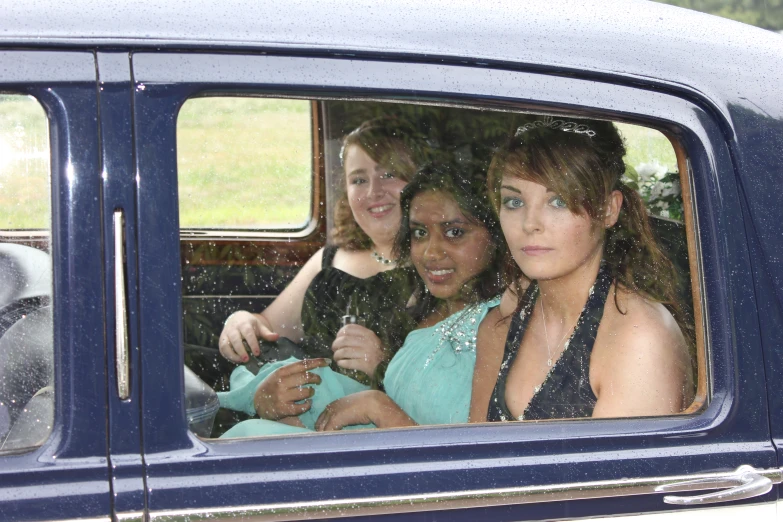three women in the back seat of a blue car, looking at the camera
