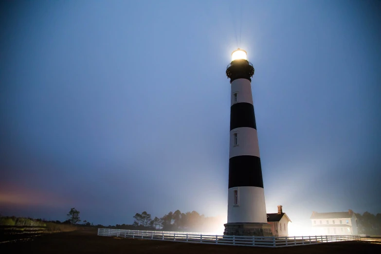 a lighthouse stands on the shore with fog rolling in