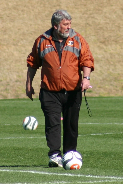 a man in an orange jacket and black pants is kicking a soccer ball on a field