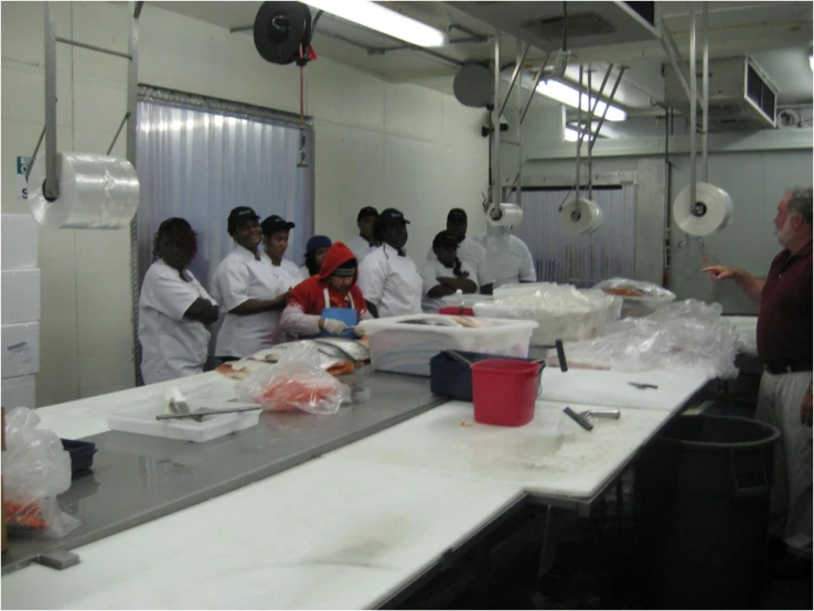 several people in white coats stand at a table making pizza