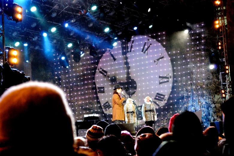 a crowd watches as two performers stand on stage with a giant clock in the background
