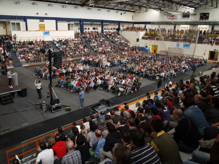 a large audience is sitting in a gym with a man speaking