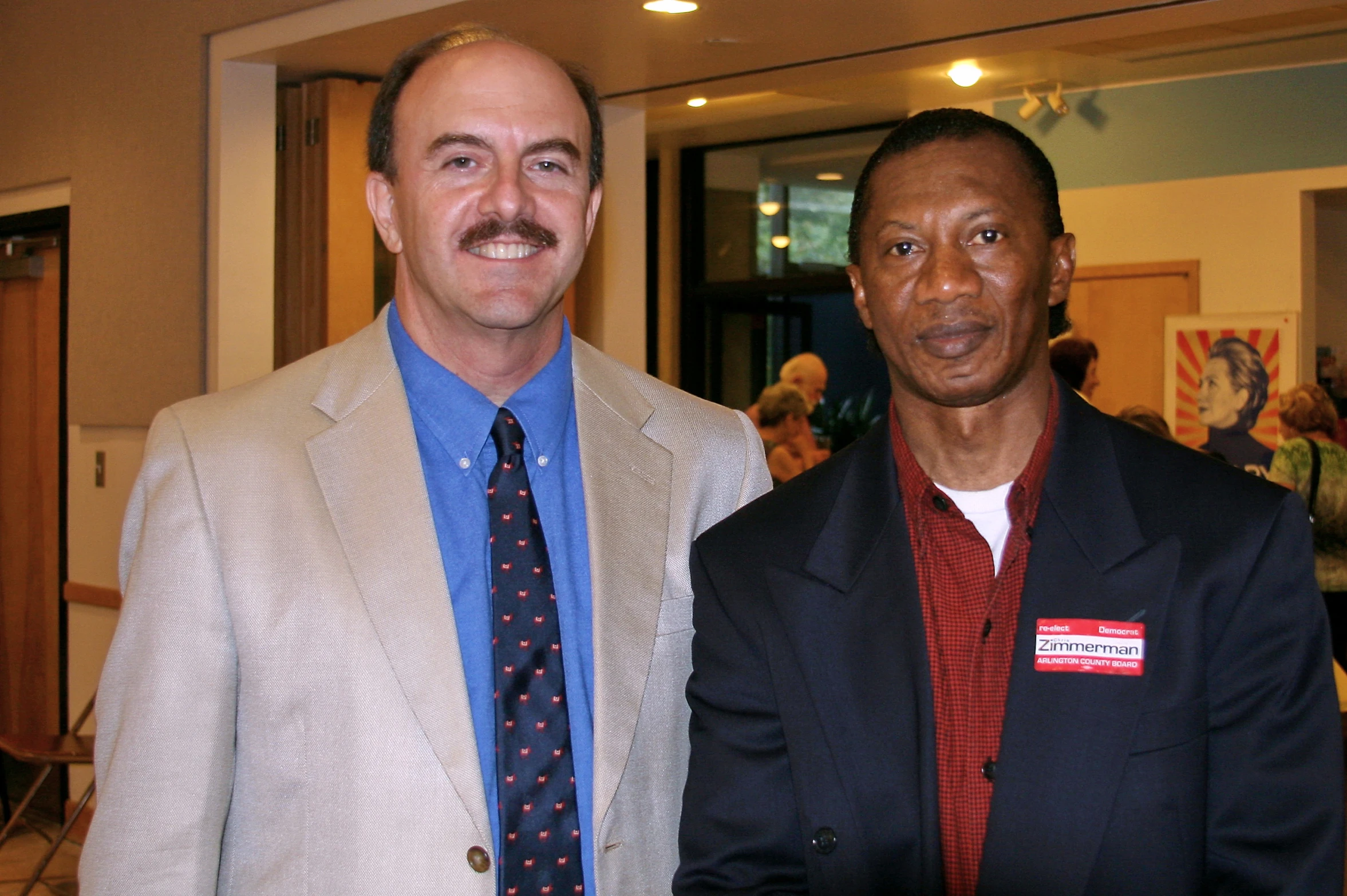 two men standing in front of a window and one is wearing a jacket and tie