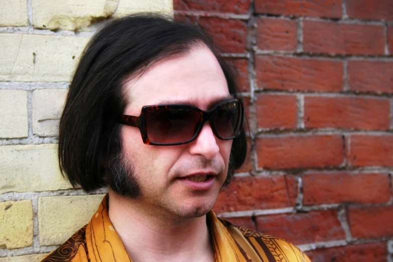 a man with sunglasses and no makeup in front of a brick wall